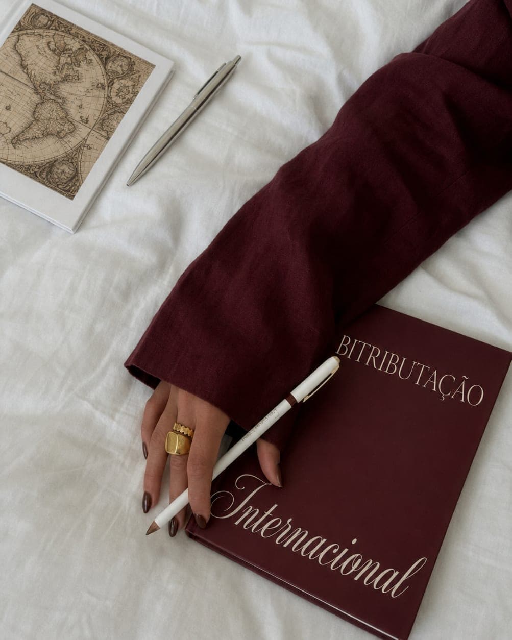 Livro de capa vinho com o título Bitributação Internacional, sobre tecido claro, ao lado de um mapa-múndi impresso, uma caneta e a mão de quem escreve