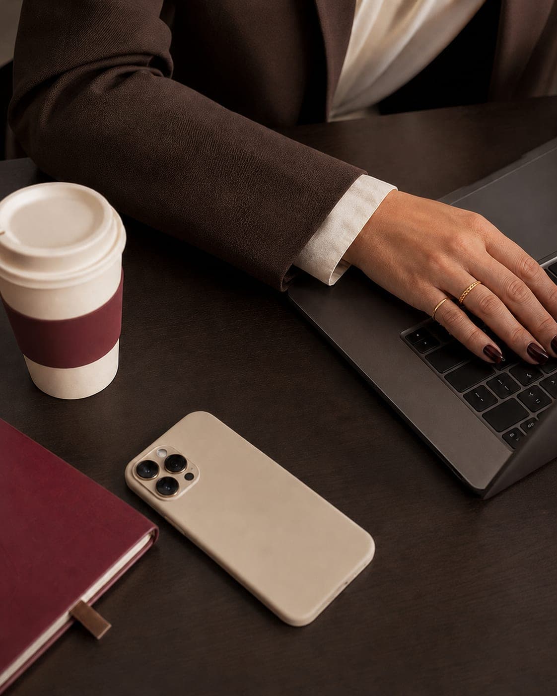 Mão sobre o teclado de um laptop, ao lado de um copo de café, um caderno vinho e um telefone